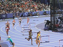 Photo of eight athletes in the bend of a purple athletics track with hurdles