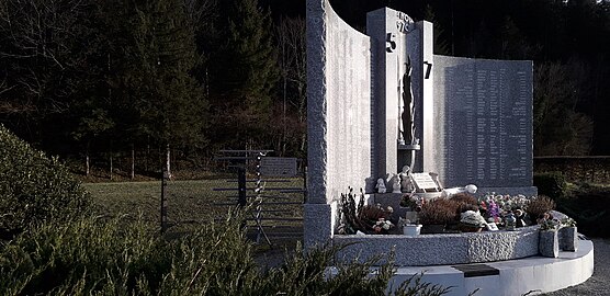 À droite, le monument aux morts en marbre ; derrière le monument, un grillage protège un tourniquet en métal.