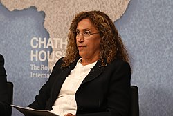 Middle Eastern woman with long hair, wearing glasses, a white top and dark jacket, sitting and looking ahead.