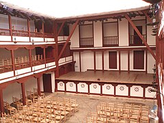 Une cour carrée à ciel ouvert à l'intérieur d'un bâtiment, deux galeries carrées de part et d'autre sont couvertes, au fond une scène surélevée puis un mur avec portes et balcons ; nombreuses chaises.