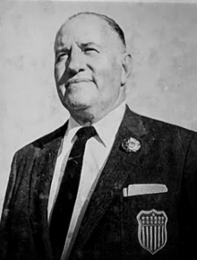 Aged balding man, wearing crisp black suit and tie, white shirt, and a large patch that almost looks like a railroad police patch, but is the patch of the U.S. Olympic Team. Nothing behind the man, possibly the sky, as he looks into the distance behind the camera, possibly watching a sports match.