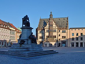 Im Vordergrund eine im Schatten liegende auf einem steinernen Podest stehende Statue. Am Podest sind zwei weitere Statuen platziert. Dahinter steht neben anderen, von der Sonne beschienen Häusern zentral ein mit Wappen verziertes fachwerkartiges Gebäude mit einem zentralen Turm und einer Uhr.