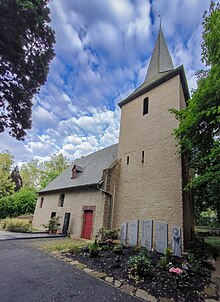 Stark untersichtiges Foto einer größeren Kapelle mit Turm und Spitzdach, drumherum gepflegte Blumenbeete, Bäume. Blauer, bewölkter Abendhimmel