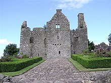 A ruined stone castle wall against blue sky, with manicured hedges in the foreground.