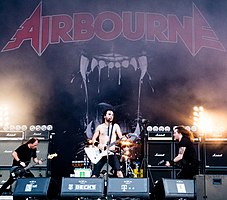 Three members of the band are in view. The band's name 'Airbourne' is displayed on a back-board, above the drummer, who is obscured behind the lead singer.