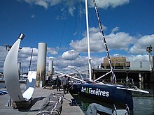 Voilier noir amarré au ponton. Des hommes mettent en place le foil tribord. Au premier plan, le foil bâbord est posé sur le ponton.