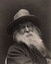 A smiling old man with a hat, dark jacket and long grey beard