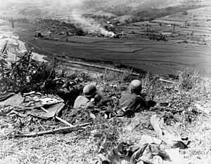 Armed men sit in foxholes watching over a lower terrain feature