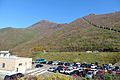 Photo couleur d'un parking comprenant plusieurs voitures stationnées au pied d'une montagne boisée, sous un ciel bleu.