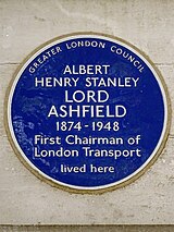 A circular blue ceramic plaque with white raise lettering fixed to a brick wall bears the text "ALBERT HENRY STANLEY LORD ASHFIELD 1874-1948 First Chairman of London Transport lived here".