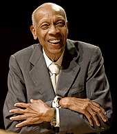 An aged man smiling dressed in a grey suit with his arms crossed.