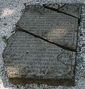 Photographie en couleurs d'une pierre tombale morcelée et gravée d'incriptions