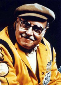 Photo of a late middle-aged man with a moustache, wearing eyeglasses, a yellow leather hockey jacket, and a Tam o' shanter cap