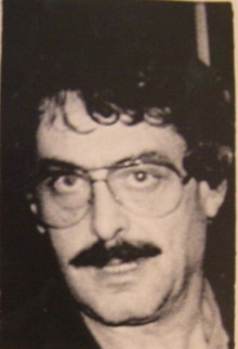A headshot of a disheveled-looking man with glasses and a mustache.