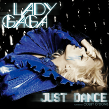 Lady Gaga laying down wearing a dark blue dress with a big, layered collar. Her right hand touches her forehead. Beneath her left eye is a blue-colored lightning-shaped sticker.