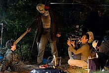 A man with a video camera kneels to film as an actor pulls a machete from the head of another actor who is sitting on the ground. Behind the cameraman, another man records sound from the scene they are filming, while a third man stands in the background.