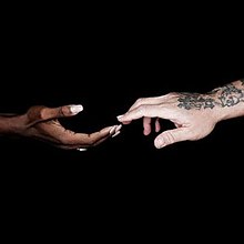 A female black hand approaches a male white hand with various tattoos on it.