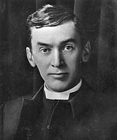 young white man, clean shaven, dark haired in clerical dress, looking severely towards the camera