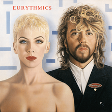In a white and blue background, Annie Lennox (on the left; seen without clothing), and Dave Stewart (seen on the right side, seen wearing a suit) stare at the viewer. The text "EURYTHMICS" appear in red on top of Annie. A oval-shaped pin with a heart hanging out at the bottom is on Dave's suit with the pin reading "Revenge".