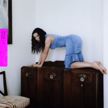 A woman with long black wavy hair and a blue dress poses for the camera on her hands and knees atop a piece of wood furniture.